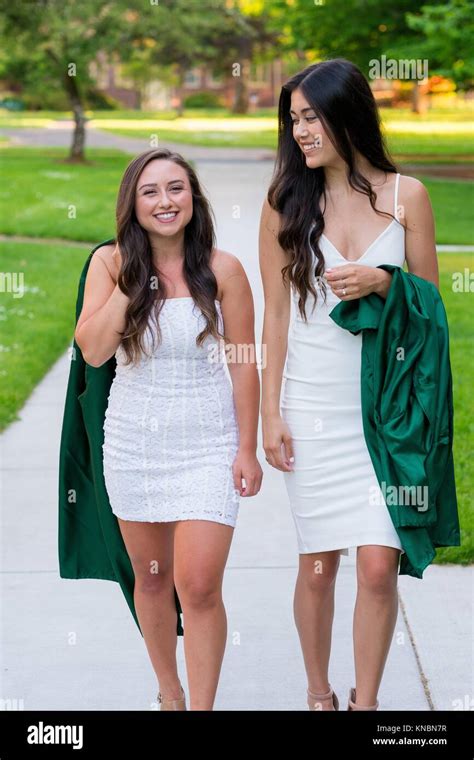 Two College Girls And Best Friends Walk On A Sidewalk While Holding Their Caps And Gowns Before