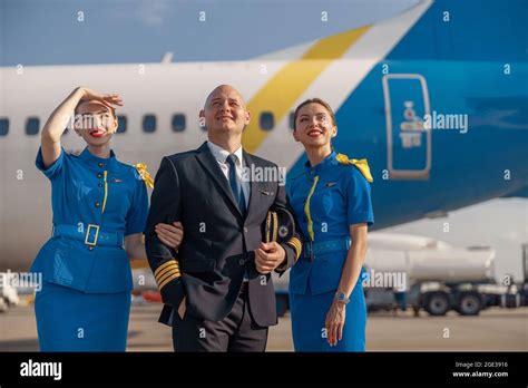 Happy Pilot And Two Pretty Stewardesses Standing Together In Front Of