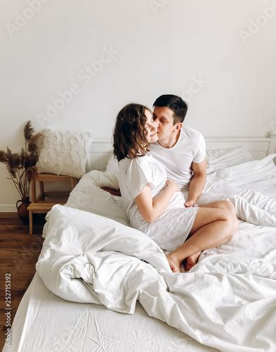 Happy Young Husband Kissing His Smiling Pregnant Wife And Hugging Belly Bump On White Bed