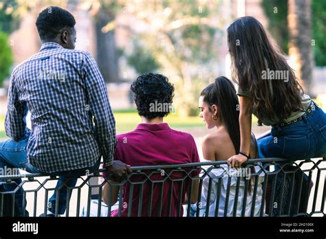 Back Of Group Of Multiethnic Friends Sitting On A Bench Of A Park While