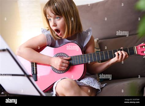 Surprised Girl Practicing The Guitar Looking At A Sheet Music And Sitting On A Sofa In Her