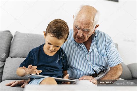 Grandfather And Grandson Sitting Together On Couch At Home And Using