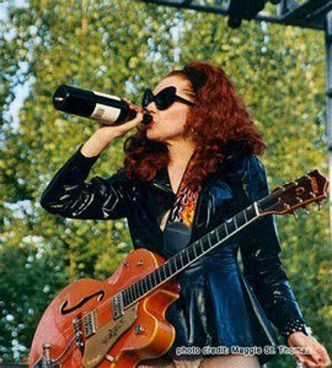 A Woman Singing Into A Microphone While Holding A Guitar In Her Right Hand And Wearing Sunglasses