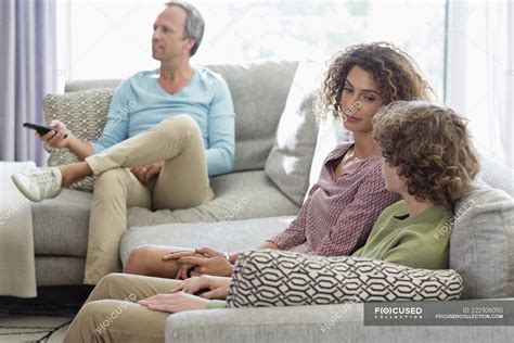Mother Talking To Son On Sofa While Father Watching Tv On Background In