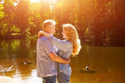 An Adult Couple Stand On The Shore Of The Lake In An Embrace In The Rays Of The Setting Sun