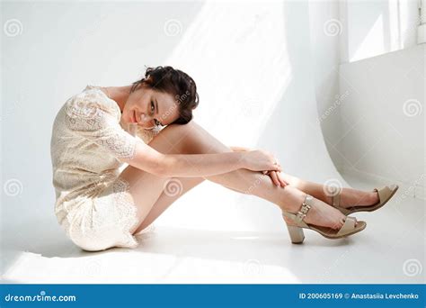 Pretty Happy Woman In White Short Dress Sitting On The Floor Hugging Her Knees And Looking In