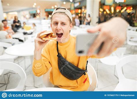 Happy Young Man Sitting At Fast Food Restaurant Eating Burger And