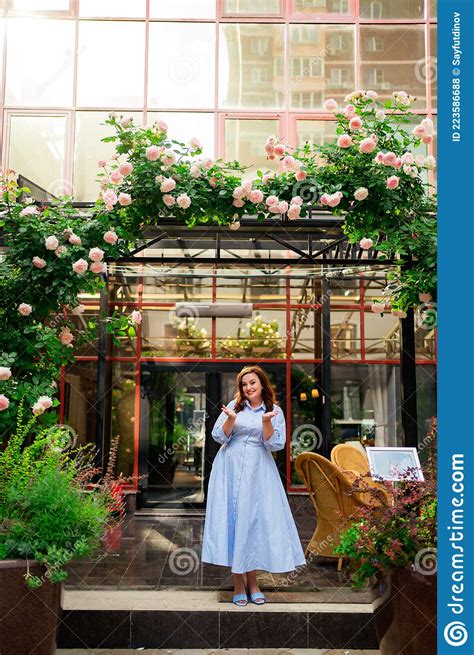 A Pretty Woman In A Blue Dress Stands In The Arch Of A Climbing Rose