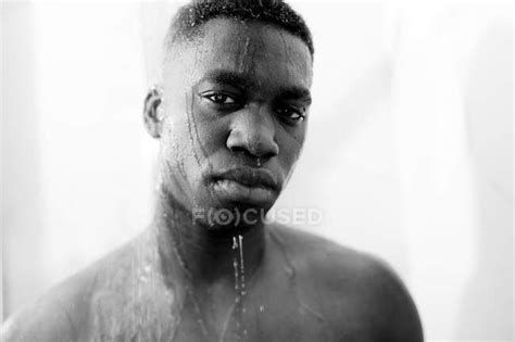 Black And White Of Emotionless Young Black Guy Taking Shower In Light Bathroom And Looking At