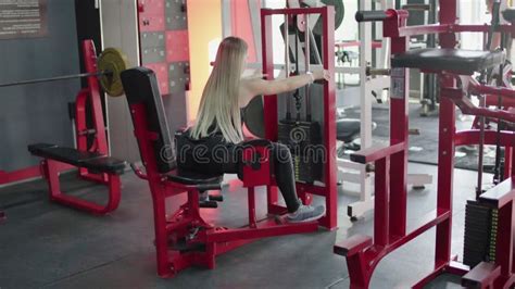 Back View Of Blonde Woman Does Squats With Barbell On Smith Machine At