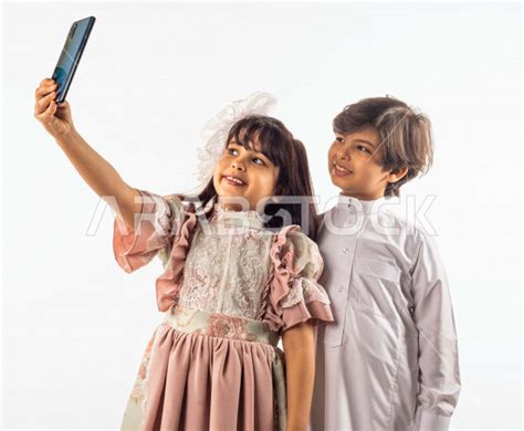 خلفية بيضاء لصبي وفتاه صغيران سعوديان مبتسمان ، تمسك الفتاه الجوال بيدها يقومان بإلتقاط صور