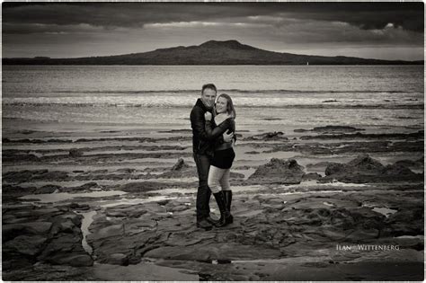 A Man And Woman Standing On Rocks Near The Ocean With An Island In The