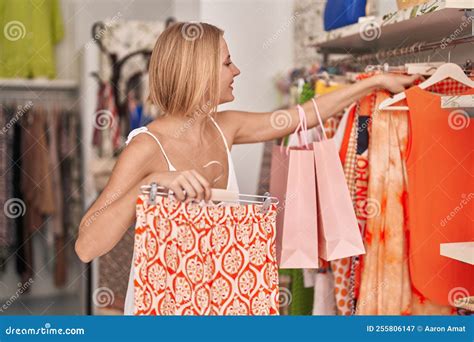 Young Blonde Woman Customer Smiling Confident Holding Clothes On Rack At Clothing Store Stock