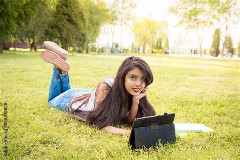 Young Indian Girl Lying In The Park On The Grass Holding A Tablet She Looks At The Camera And
