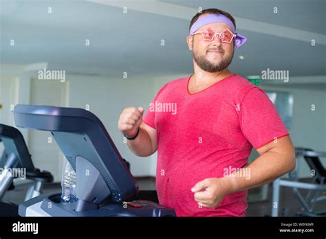 Funny Fat Male In Pink Glasses And In A Pink T Shirt Is Engaged On A Treadmill In The Gym