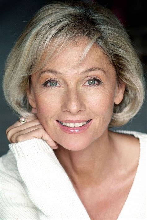 An Older Woman With Blonde Hair And Blue Eyes Smiles At The Camera While Wearing A White Sweater