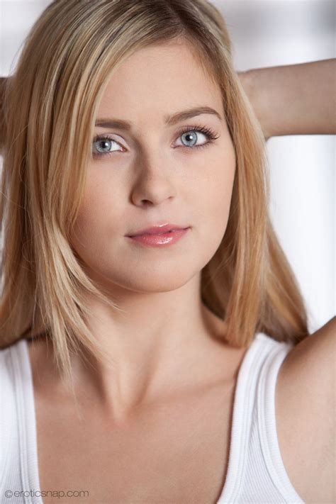 A Woman With Blonde Hair And Blue Eyes Posing For The Camera Wearing A White Tank Top