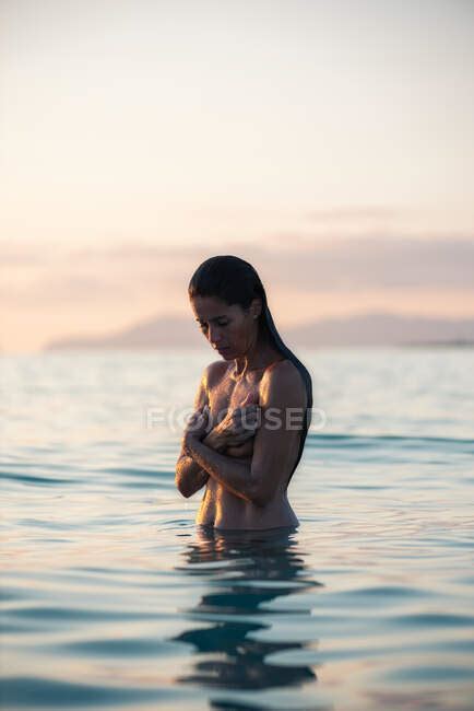 Mujer desnuda cubriendo el pecho mientras está de pie en el agua de mar