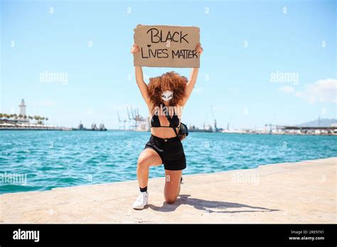 Serious Ethnic Female With Afro Hairstyle Kneeling On Pavement With Cardboard Poster With Black