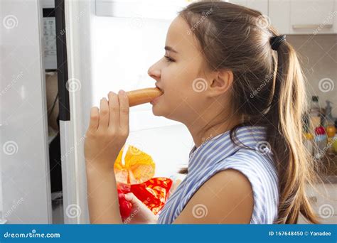 The Girl Looks In The Refrigerator Hungry Eats Sausage Stock Photo