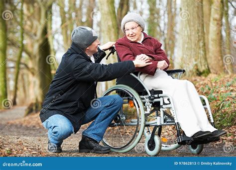 Caregiver Man Walking With Disabled Senior Woman At Wheelchair In
