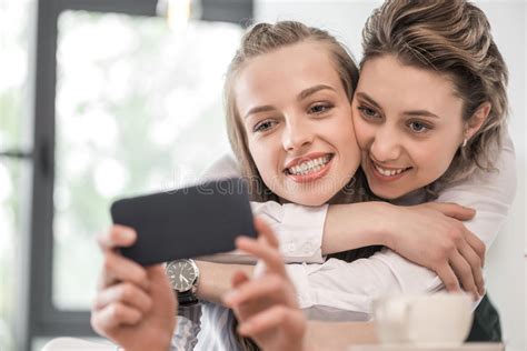 Smiling Girlfriends Embracing And Taking Selfie On Smartphone At Cafe