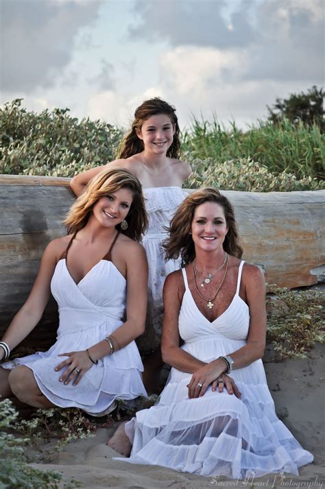 Three Women In White Dresses Are Sitting On The Beach And One Is Leaning Against A Log