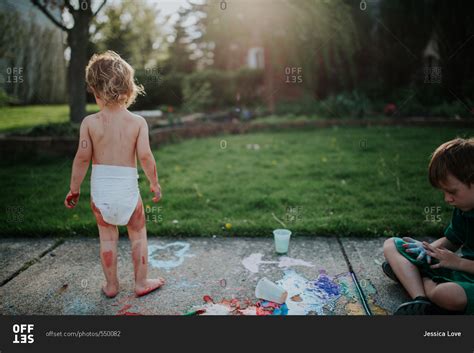 Toddler In Diaper And Brother Playing With Chalk Paint On Sidewalk