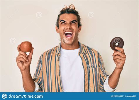 Young Hispanic Man Holding Red Apple And Donut Angry And Mad Screaming Frustrated And Furious