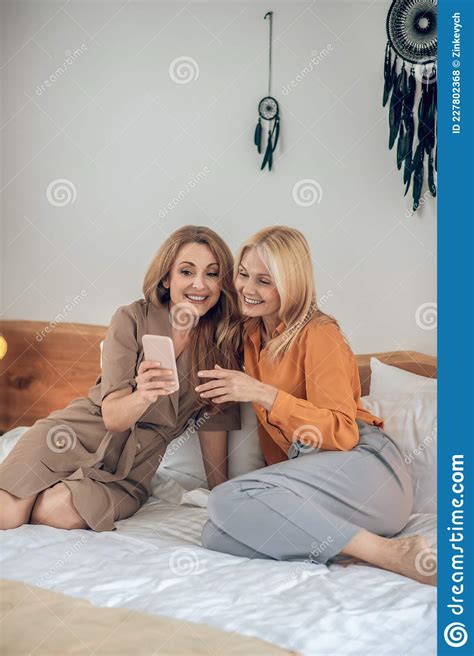 Two Smiling Women Sitting On The Bed And Looking Photos On Internet
