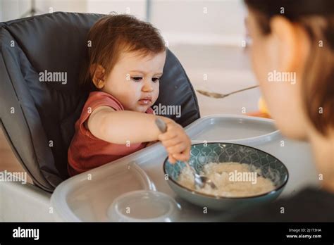 Baby Girl Sitting In A High Chair Trying To Eat Alone Porridge With