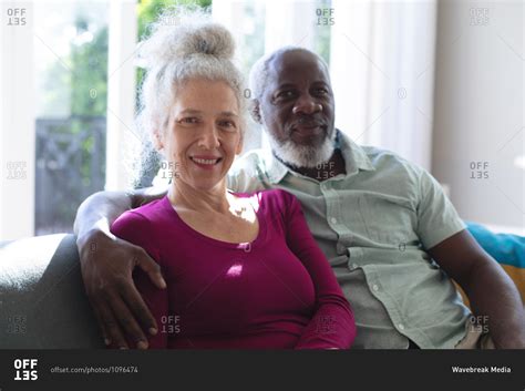 Senior Mixed Race Couple Embracing Looking At The Camera And Smiling In Living Room Staying At