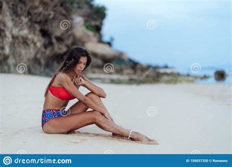 Slender Tanned Woman In Colored Swimsuit Sits On Ocean Beach With White