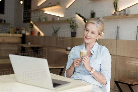 Smiling Mature Woman Drinking Coffee And Using Laptop In Cafe Stock