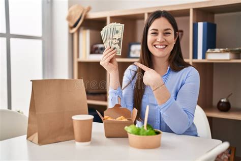 Young Brunette Woman Eating Take Away Food At Home Holding Money