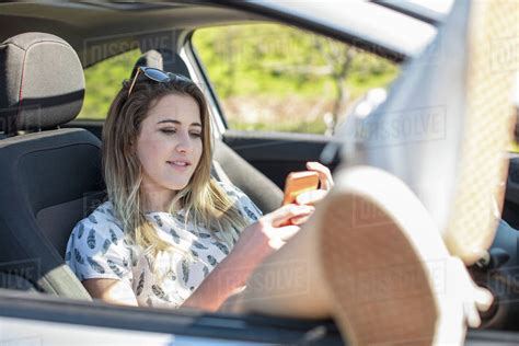 Young Woman Sitting In Car Using Smartphone Legs Out Of Open Car