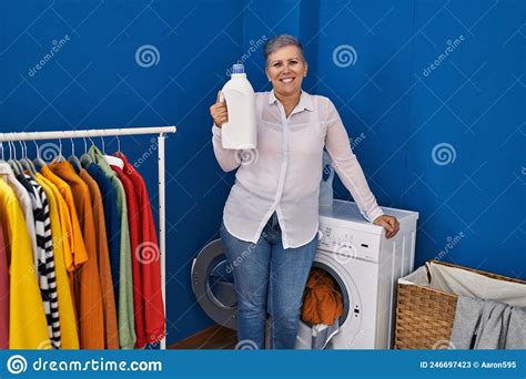Middle Age Woman Smiling Confident Holding Detergent Bottle At Laundry