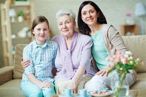 Grandmother With Her Daughter And Granddaughter Sitting On A Couch Smiling At The Camera Stock