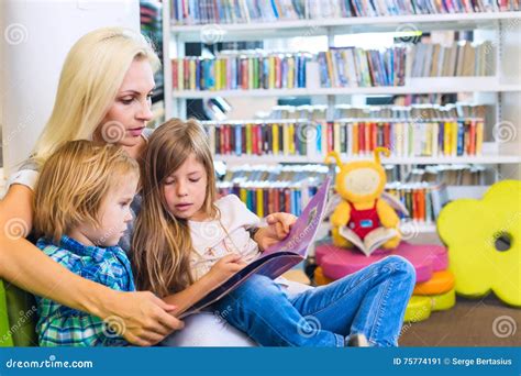 Mother With Little Girl And Boy Read Book Together In Lounge Stock