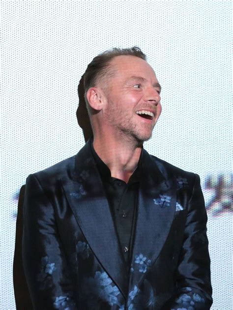 A Man In A Black Suit Laughing While Standing Next To A White Wall With