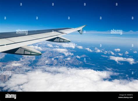 Airplane Window View Showing Wing Of Plane Flying Over Clouds And Rocky