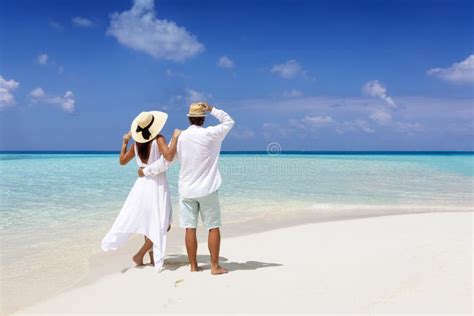 A Beautiful Tourist Couple With Sunhats Stands On A Tropical Beach