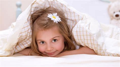 Cute Little Girl Is Lying Down On White Bed Covered With White Bedsheet