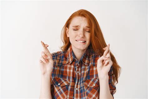 worried redhead girl making a wish with crossed fingers and closed eyes biting lip nervously