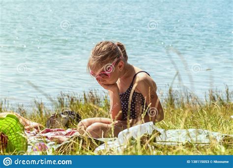 A Girl In A Bathing Suit Sits Lonely On The Beach On The Grass Near The River On A Sunny Day