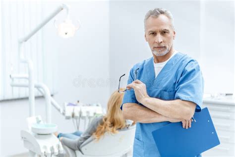 Dentist In Uniform With Clipboard In Dental Clinic With Patient Behind