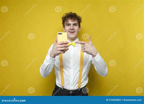 Young Guy In Bow Tie And Suspenders Holds Smartphone And Points To It With Finger On Yellow