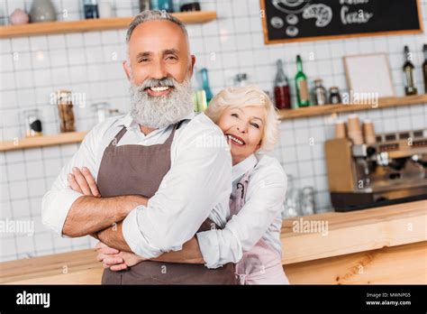 Portrait Of Smiling Senior Woman Hugging Husband In Apron In Coffee
