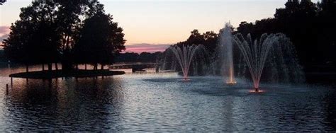 Several Fountains In The Middle Of A Lake At Sunset With Trees And