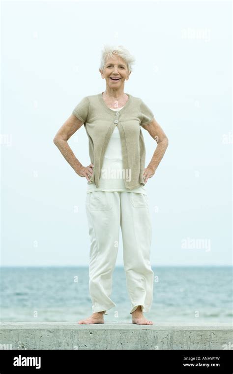 Senior Woman Standing With Hands On Hips Smiling At Camera Ocean In Background Full Length